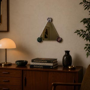 May include: A triangular mirror with decorative pom-poms at each corner. The mirror is hanging on a beige wall. A wooden cabinet with books, a lamp, and a vase is below the mirror. The books are titled "Francis Bacon", "Lucian Freud Paintings", and "Picasso".