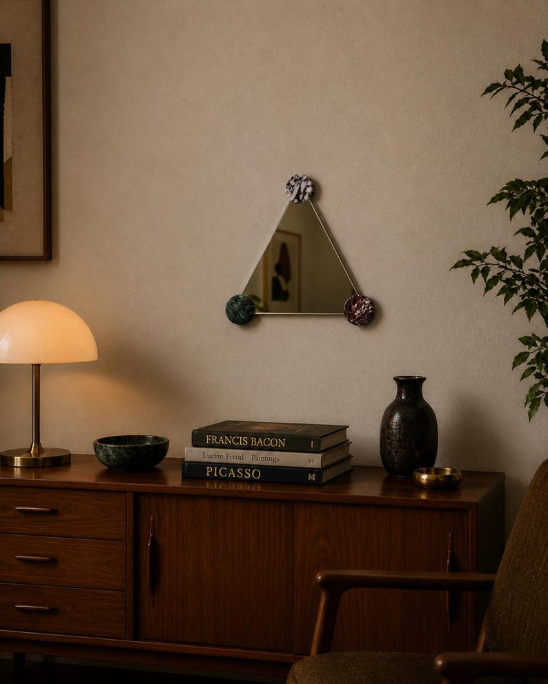 May include: A triangular mirror with decorative pom-poms at each corner. The mirror is hanging on a beige wall. A wooden cabinet with books, a lamp, and a vase is below the mirror. The books are titled "Francis Bacon", "Lucian Freud Paintings", and "Picasso".