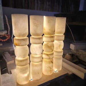 May include: Four cylindrical lamps made of translucent white stone with natural beige veining. Each lamp features a stacked design with alternating smooth cylindrical and rounded bulbous sections, emitting a warm internal glow from within.
