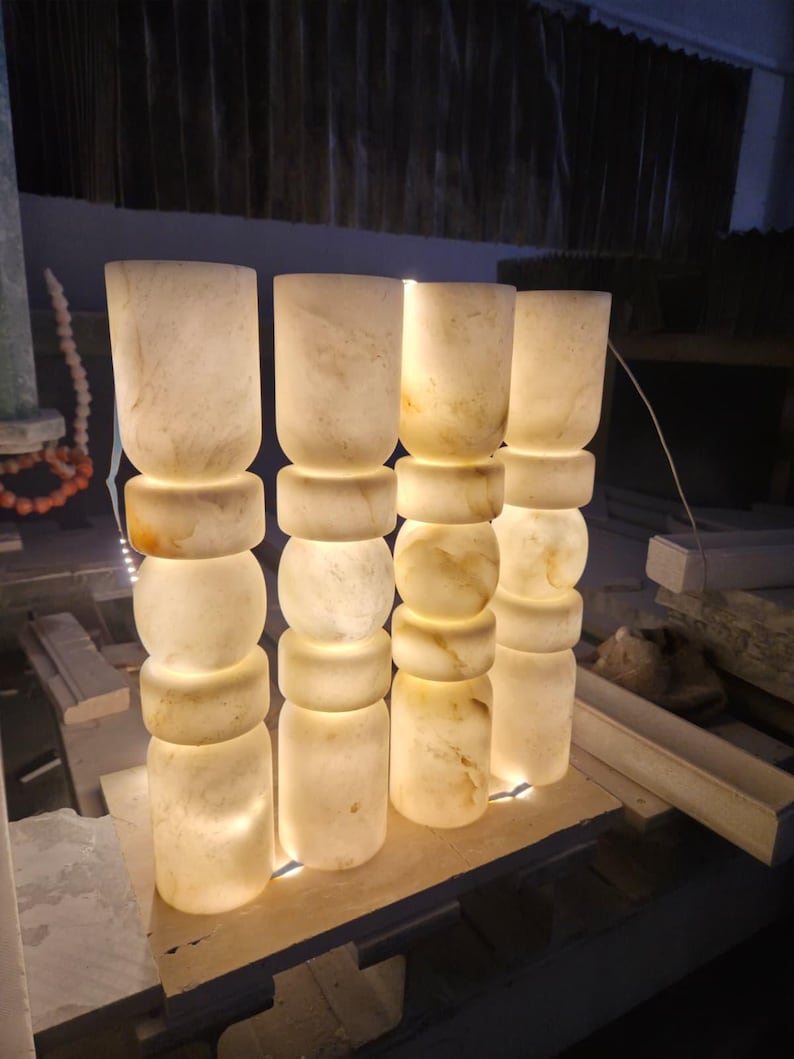 May include: Four cylindrical lamps made of translucent white stone with natural beige veining. Each lamp features a stacked design with alternating smooth cylindrical and rounded bulbous sections, emitting a warm internal glow from within.