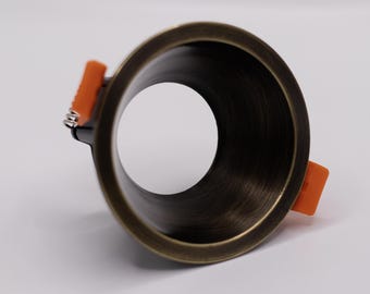 Könnte beinhalten: Eine kreisförmige Einbauleuchtenblende mit dunkelbronzefarbener, gebürsteter Metalloberfläche. Die Leuchte verfügt über eine breite, hohle Mitte und zwei leuchtend orangefarbene, federbelastete Kunststoff-Montageclips an den Seiten.