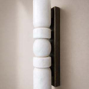 May include: A wall sconce featuring a vertical arrangement of five white marble cylindrical and spherical shapes attached to a rectangular dark bronze metal backplate. The light fixture has a stacked, geometric design with smooth surfaces.
