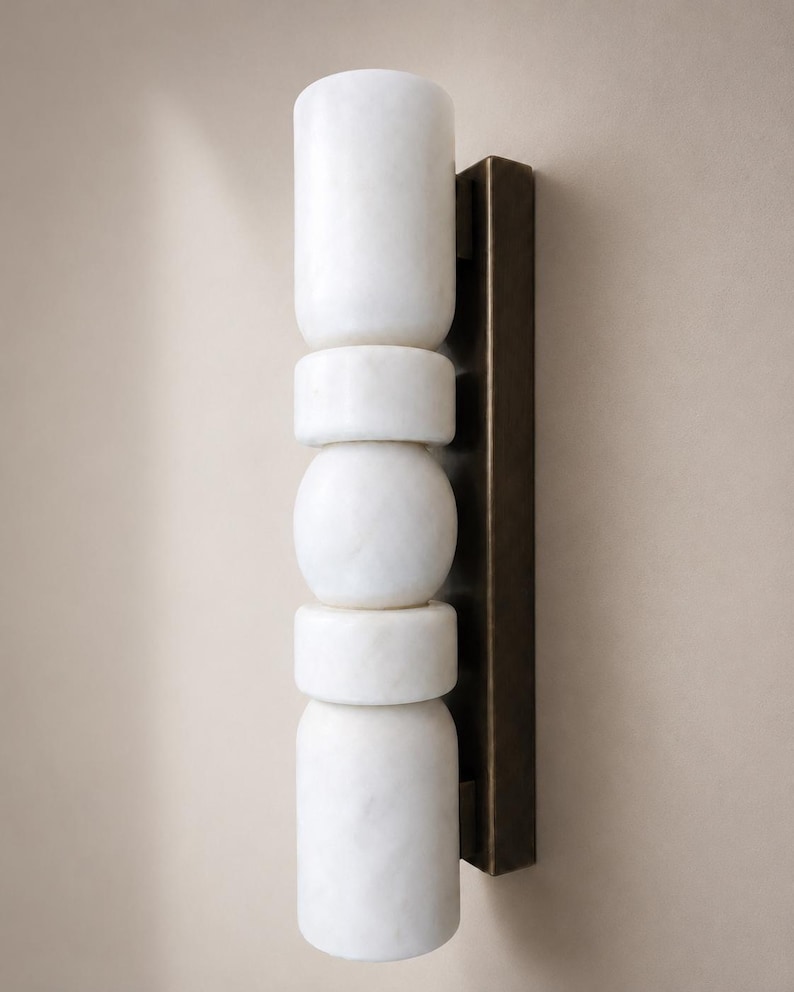 May include: A wall sconce featuring a vertical arrangement of five white marble cylindrical and spherical shapes attached to a rectangular dark bronze metal backplate. The light fixture has a stacked, geometric design with smooth surfaces.