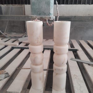 May include: Two tall, cylindrical stone pillars with a pale, off-white color and subtle beige veining. Each pillar features a stacked design consisting of a smooth top section, two rounded middle rings, and a solid base, all carved from stone.