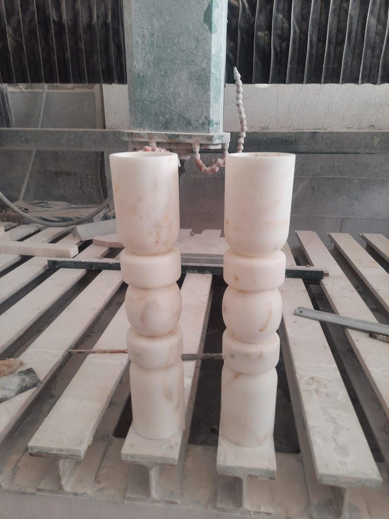 May include: Two tall, cylindrical stone pillars with a pale, off-white color and subtle beige veining. Each pillar features a stacked design consisting of a smooth top section, two rounded middle rings, and a solid base, all carved from stone.