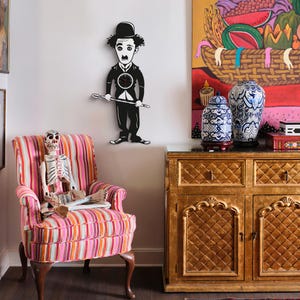 Op de afbeelding: Een zwart-witte wandklok in de vorm van Charlie Chaplin, met een wandelstok. Een kleurrijke gestreepte fauteuil met een skelet erin. Een gouden kast met twee blauw-witte vazen en een schilderij van een fruitmand.