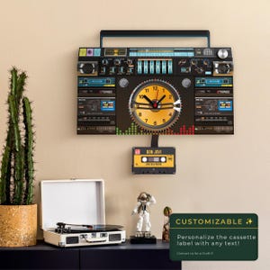 Puede incluir: Un reloj de pared con forma de radiocasete vintage con una esfera amarilla. El reloj tiene un asa negra y una etiqueta de cinta de cassette que dice "BON JOVI". El texto "CUSTOMIZABLE" está en un rectángulo verde.
