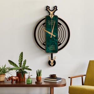 May include: A decorative wall clock with a black and gold frame, featuring a green marble-like center with gold clock hands. The clock has a deer antler design at the top and bottom, and is hanging on a white wall.