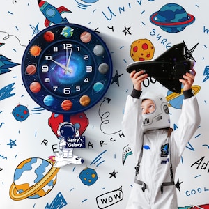 Può includere: Un orologio da parete a tema spaziale, con un design blu e nero raffigurante pianeti e una galassia. L'orologio ha un quadrante bianco con numeri e lancette nere. Un bambino con una tuta da astronauta bianca tiene in mano un'astronave giocattolo nera.