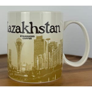 May include: A cream-coloured ceramic mug featuring the word Kazakhstan in large black letters at the top. Below the text, the Starbucks Coffee logo is printed, followed by a golden-brown illustration of the Astana city skyline and the Bayterek tower.