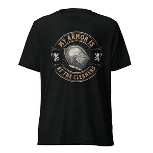 May include: Dark heather gray t-shirt with a circular graphic on the back. The graphic features a medieval helmet and the text "MY ARMOR IS AT THE CLEANERS" in a vintage style. Two heraldic lions flank the helmet design.