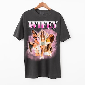 May include: Dark gray t-shirt with a graphic design featuring the word "WIFFY" in pink and white, along with images of women. The shirt is hanging on a wooden hanger.