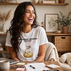 Könnte beinhalten: Ein weißes kurzärmeliges T-Shirt mit einer Grafik eines Französischen Bulldoggen, der auf einer Kartoffel sitzt. Der Text über der Grafik lautet "EMOTIONAL SUPPORT POTATO" in fetten schwarzen Buchstaben, mit "FRENCHIE MOM" darunter.