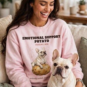 Może przedstawiać: Jasnoróżowa bluza z okrągłym dekoltem z ciemnobrązowym napisem "EMOTIONAL SUPPORT POTATO" i "FRENCHIE MOM" nad ilustracją buldoga francuskiego siedzącego na ziemniaku, z napisem "emotionally available, horizontally" poniżej.