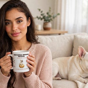 Könnte beinhalten: Ein weißer Keramikbecher mit einer Grafik einer Französischen Bulldogge, die auf einer Kartoffel sitzt. Der Text EMOTIONAL SUPPORT POTATO und FRENCHIE MOM ist zusammen mit kleinen roten Herzillustrationen auf der Vorderseite zu sehen.