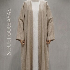 May include: A beige open-front abaya with long sleeves worn over a plain white floor-length dress. The garment features a textured fabric and the text SOUËIRAABAYAS is printed vertically in white on the left side of the display.