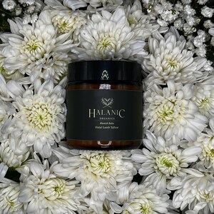 May include: A white jar with a black lid and label. The label says "HALANIC ORGANICS Blemish Balm Halal Lamb Tallow". The jar is surrounded by white flowers.