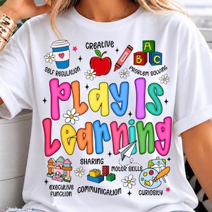 May include: A white t-shirt featuring the colourful text Play Is Learning. The design includes illustrations of a coffee cup, apple, crayon, alphabet blocks, paper airplane, building blocks, and a paint palette, with labels like Self Regulation.