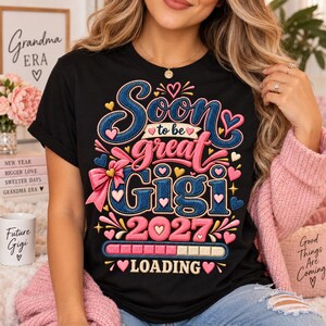 May include: A black t-shirt featuring a graphic design with the text Soon to be great Gigi 2027 LOADING. The design includes blue and pink lettering, a pink bow, and various heart shapes, with a loading bar graphic in pink and gold colors.