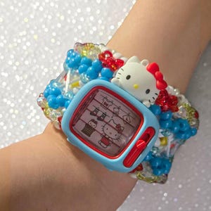 May include: A beaded bracelet featuring a light blue television-shaped frame with a red border. The screen displays a graphic of Hello Kitty. A white Hello Kitty figure with a red bow is attached to the top. The band consists of blue and clear beads.