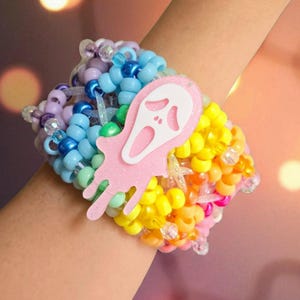 Pode incluir: Uma pulseira de contas coloridas com um pingente de rosto de fantasma rosa e branco. A pulseira apresenta um arco-íris de contas em tons de azul, amarelo, laranja, rosa, verde e roxo. O pingente é rosa claro com um rosto de fantasma branco.