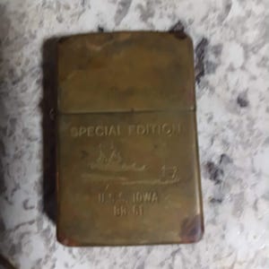 May include: A rectangular, brass-colored lighter with the text "SPECIAL EDITION" and an image of a ship. Below the image is the text "U.S.S. IOWA BB 61". The lighter has a worn, vintage appearance.