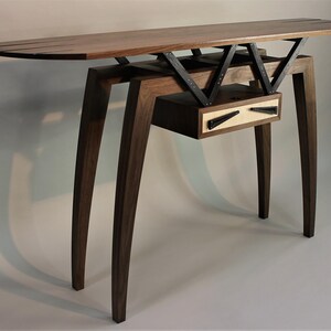 May include: A modern console table with a walnut wood top and legs. The table features a drawer with a black metal handle and a unique geometric design in the base.