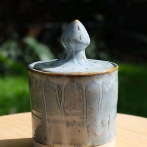 May include: A handmade ceramic jar with a lid. The jar is cylindrical with a light blue glaze and a white base. The lid has a pointed finial. The jar is approximately 13 cm tall.