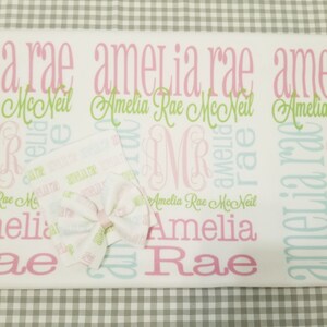 May include: A white baby blanket with a pink and green pattern. The blanket is personalized with the name "Amelia Rae" repeated multiple times. A matching hat and bow are also included.