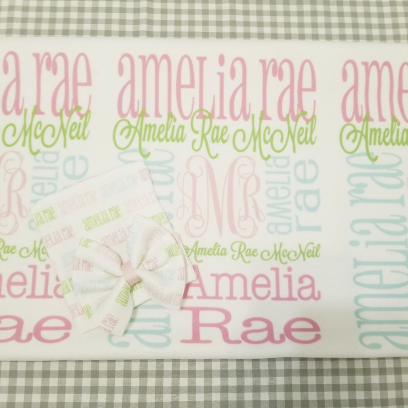 May include: A white baby blanket with a pink and green pattern. The blanket is personalized with the name "Amelia Rae" repeated multiple times. A matching hat and bow are also included.