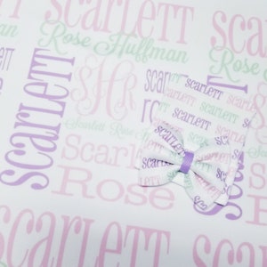 May include: A white fabric with a purple and pink pattern of the name "Scarlett" repeated. A purple bow is centered on the fabric.