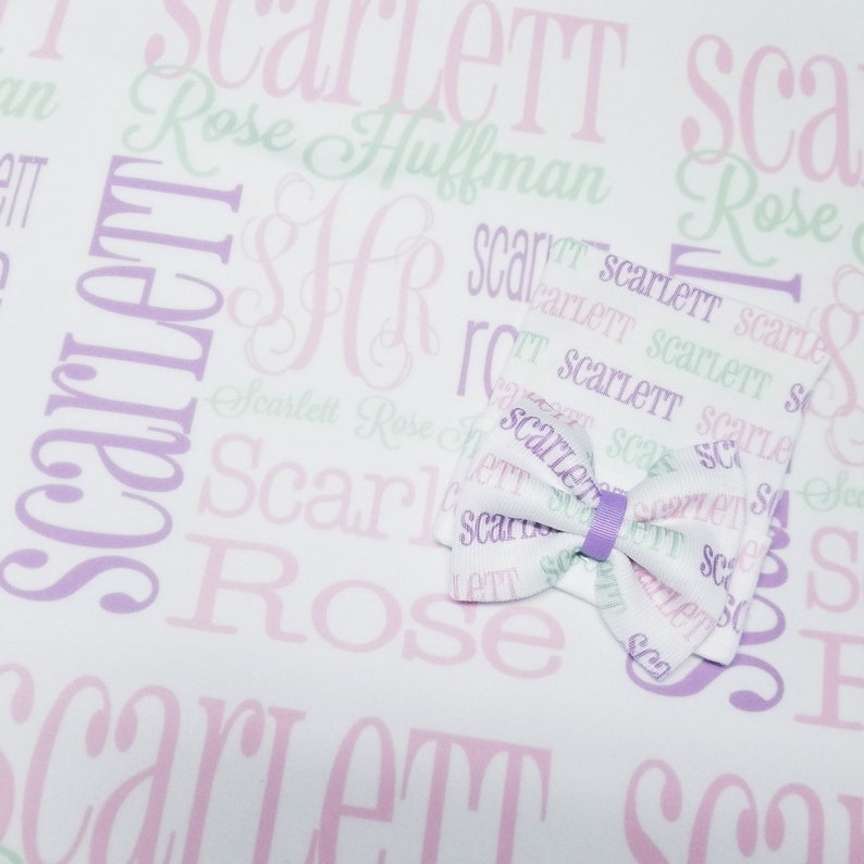May include: A white fabric with a purple and pink pattern of the name "Scarlett" repeated. A purple bow is centered on the fabric.