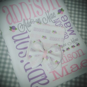 May include: A white baby blanket with the name "Addison Mae" printed in pink and purple lettering. The blanket is decorated with small purple flowers. A matching white burp cloth with a pink bow is also included.