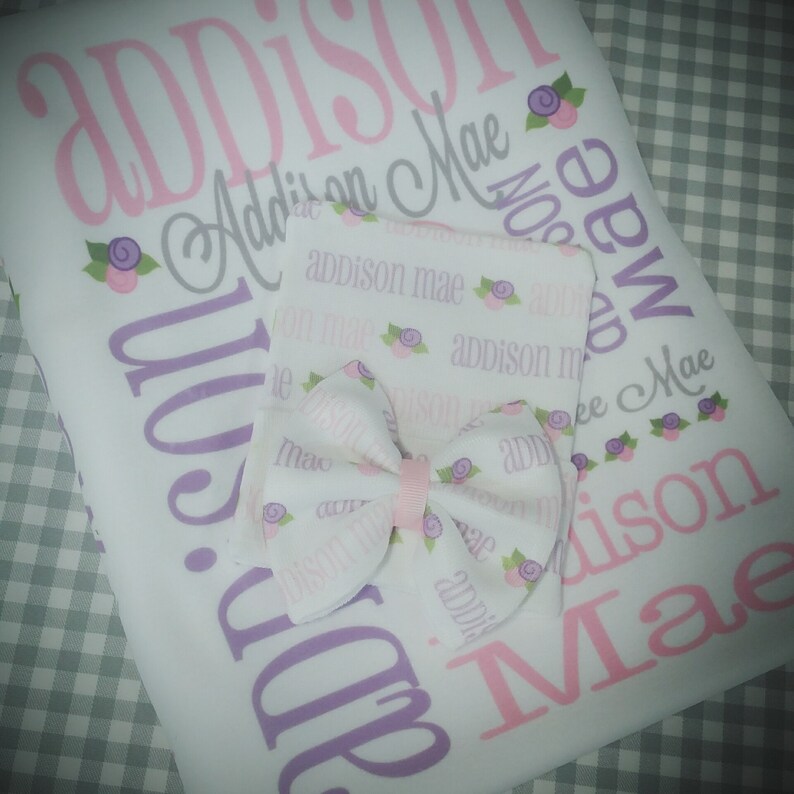May include: A white baby blanket with the name "Addison Mae" printed in pink and purple lettering. The blanket is decorated with small purple flowers. A matching white burp cloth with a pink bow is also included.