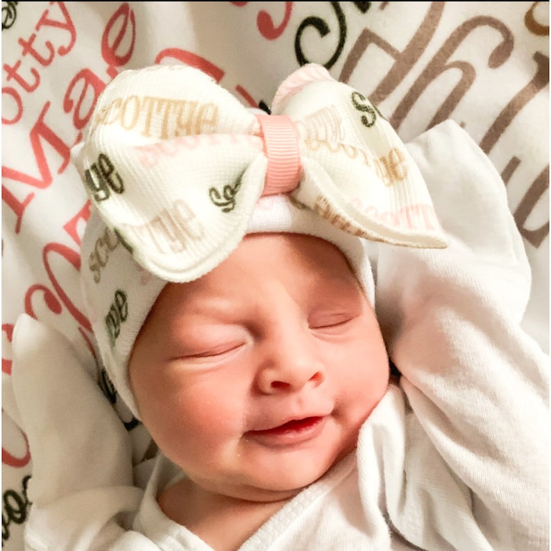 May include: A newborn baby wearing a white and pink patterned hat with a large bow. The hat has the words "Sweet Dreams" printed on it.