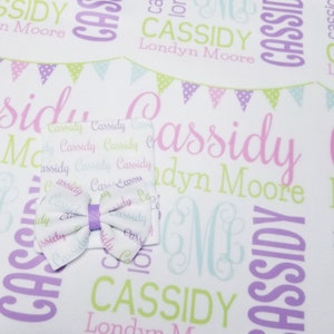 May include: A white baby blanket with a purple, green, and pink pattern of flags and the name "Cassidy Londyn Moore" repeated throughout. A white hat with the same pattern and a purple bow is on top of the blanket.