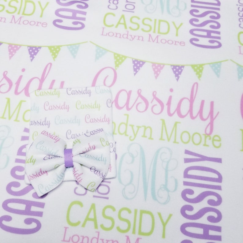 May include: A white baby blanket with a purple, green, and pink pattern of flags and the name "Cassidy Londyn Moore" repeated throughout. A white hat with the same pattern and a purple bow is on top of the blanket.