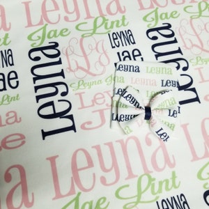 May include: A white baby blanket with a repeating pattern of the names "Leyna" and "Jae" in pink and green lettering. A navy blue bow is centered on the blanket.