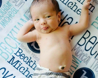 May include: A newborn baby with fair skin, wearing grey and white striped trousers, lies on a white blanket with blue text. The baby has its arms raised and is sticking out its tongue. The blanket has the name "Beckham Micheal" printed on it.