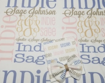 May include: A white baby blanket with a repeating pattern of the names "Indie" and "Sage" in blue, pink, and gold lettering. The blanket is paired with a matching white baby hat with a bow.