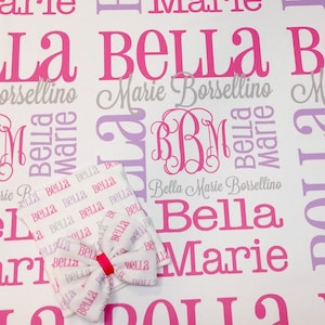 Puede incluir: Una manta de beb&eacute; blanca con un patr&oacute;n rosa y morado que presenta los nombres "Bella Marie Borsellino" repetidos en toda la superficie. La manta tiene un lazo rosa y un monograma "BBM" en el centro.