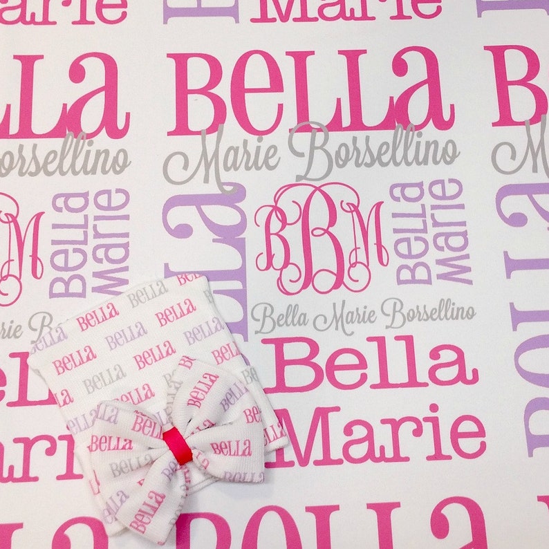 Puede incluir: Una manta de beb&eacute; blanca con un patr&oacute;n rosa y morado que presenta los nombres "Bella Marie Borsellino" repetidos en toda la superficie. La manta tiene un lazo rosa y un monograma "BBM" en el centro.