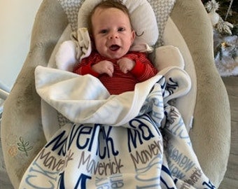 May include: A baby in a red striped top sits in a beige and white baby swing with a bunny-shaped headrest. A white and blue blanket with the name "Maverick" is draped over the baby.