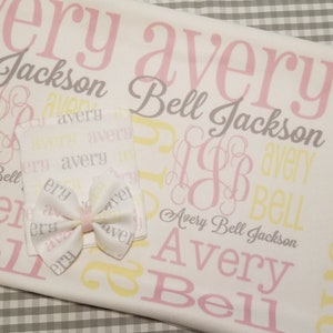 May include: A white baby blanket with a pink and yellow pattern. The blanket has the name "Avery Bell Jackson" written on it in a cursive font. There is a matching hat and bow with the same pattern.