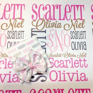 Puede incluir: Una manta de beb&eacute; blanca con los nombres Scarlett, Olivia y Niel impresos en rosa, marr&oacute;n y gris. La manta est&aacute; decorada con un lazo rosa y blanco.