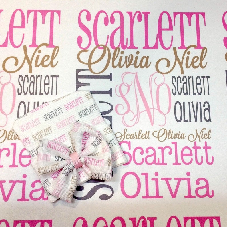 Puede incluir: Una manta de beb&eacute; blanca con los nombres Scarlett, Olivia y Niel impresos en rosa, marr&oacute;n y gris. La manta est&aacute; decorada con un lazo rosa y blanco.