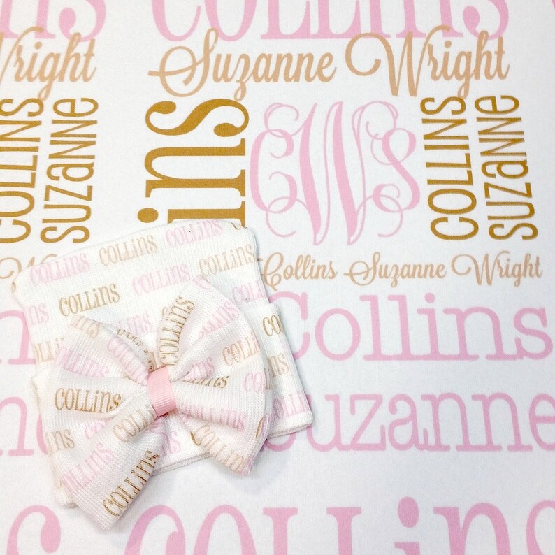 Puede incluir: Un gorro de beb&eacute; blanco con un lazo rosa y letras doradas que dicen "Collins" repetidas en el gorro. El gorro est&aacute; sobre una manta blanca con letras rosas y doradas que dicen "Collins Suzanne Wright".