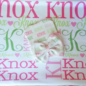 May include: A white baby blanket with a pink and green pattern featuring the name "Knox" repeated throughout. A matching white hat with a pink bow is on top of the blanket.