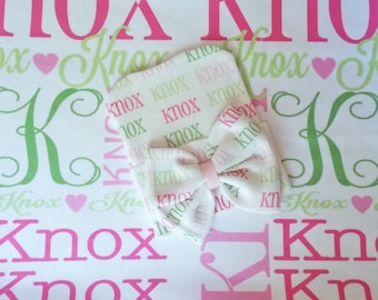 May include: A white baby blanket with a pink and green pattern featuring the name "Knox" repeated throughout. A matching white hat with a pink bow is on top of the blanket.