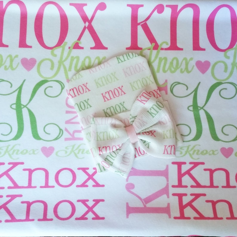 May include: A white baby blanket with a pink and green pattern featuring the name "Knox" repeated throughout. A matching white hat with a pink bow is on top of the blanket.